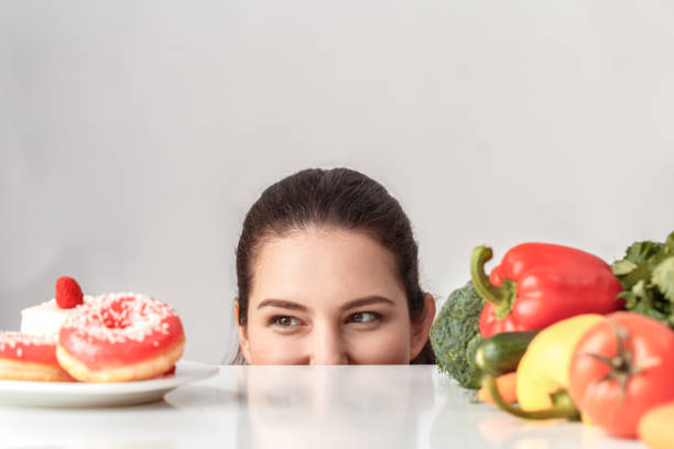 istock Body Care. Chubby girl standing hiding under kitchen table with vegetables looking at desserts smiling playful close-up 1188510248