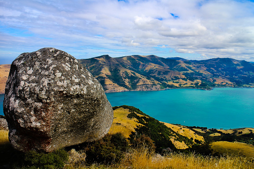 View from the top of the volcanic hills of Akaroa with a boulder in the foreground onto Akaroa Bay, an inlet of the Pacific ocean on the Banks Peninsula, Canterbury, New Zealand.