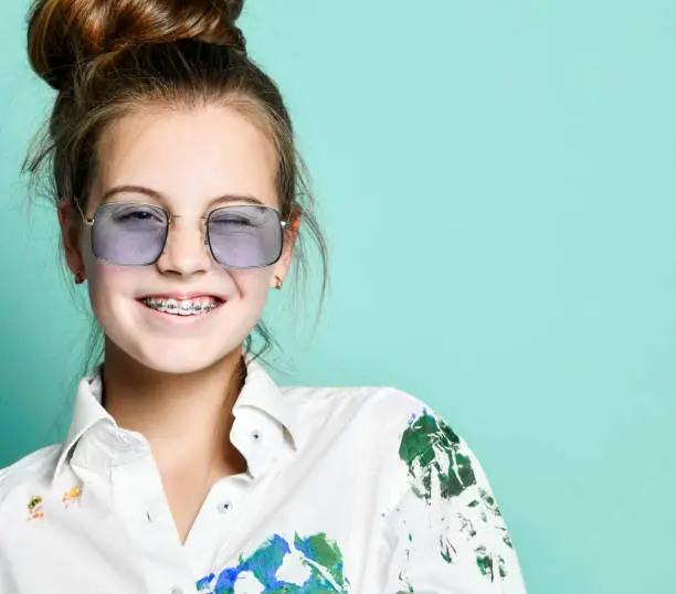 Portrait of smiling young woman girl with braces in white shirt with colorful paint stains and rectangular translucent glasses is frolic winking on gray background with copy space