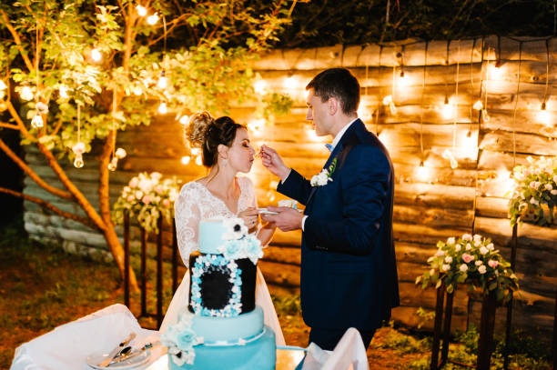 istock Bride and a groom is cutting, eating rustic wedding cake outdoors. Hands cut the cake with delicate blue and white flowers. Couple in magical evening forest decorated light garlands. Night ceremony. 1188549502