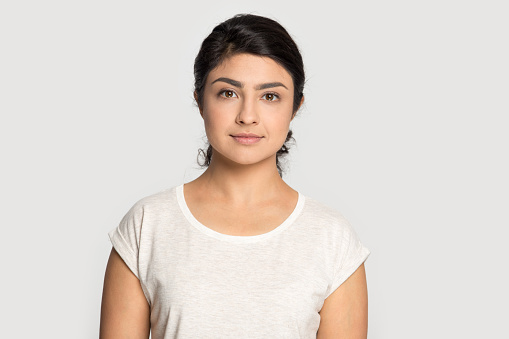 Head shot portrait close up beautiful Indian girl with perfect skin posing isolated on grey studio background, pretty young woman wearing white t-shirt looking at camera, natural makeup