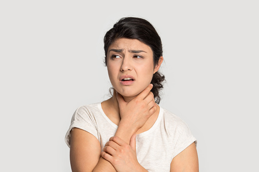 Sick upset Indian girl touching neck, suffering from sore throat, standing isolated on grey studio background, unhappy young woman feeling bad, unwell, painful swallowing, illness concept