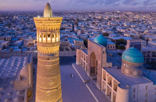 Aerial view of illuminated Kalyan Minaret at Po-i-Kalyan - Poi Kalan during colorful sunset twilight over the famous old town in the city of Bukhara - Buxoro - Бухорo. Iconic Kalyan Minaret in the foreground, Miri Arab Madressa on the right. Unrecognizable wedding couple - bride and groom in the middle of the Poi Kalan Compex. Drone Point of View. Itchan Kala, Bukhara - Бухорo, Khorezm Region, Uzbekistan, Central Asia.