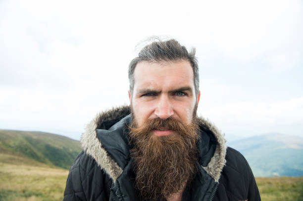 istock Man with long beard and mustache wears jacket. Hipster on strict face with beard looks brutally while hiking. Hermit concept. Man with brutal bearded appearance, brutal unshaven man looks untidy 1188570546