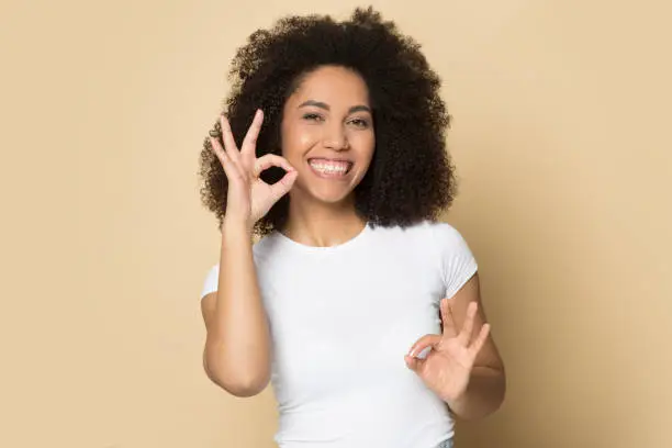 Head shot portrait close up smiling African American girl speaking sign language, standing isolated on brown background, deaf speechless young female communicating, looking at camera