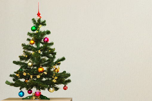Artificial green Christmas tree with colorful Christmas toys, standing on a table against a gray wall.