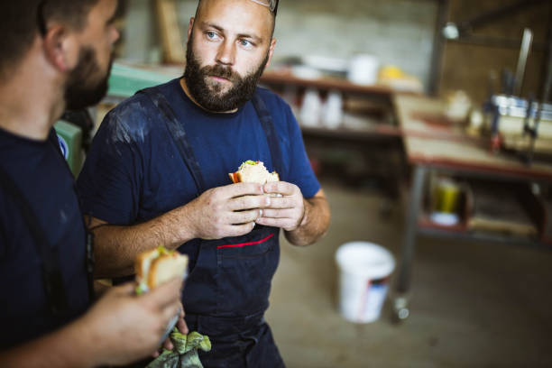 Young manual workers talking while eating sandwiches on a break in a workshop. Young manual workers communicating while eating sandwiches on a lunch break in a workshop. construction lunch break stock pictures, royalty-free photos & images