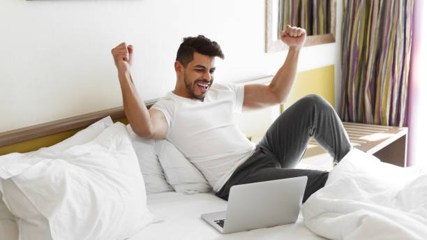 istock Young man celebrating success, sitting on bed, using laptop 1188588780