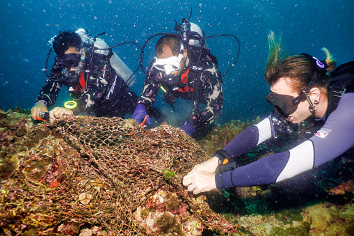 Ko Rok, Andaman sea, Krabi province, Thailand - October 10 2019:  Three male scuba divers are cutting discarded fishing net from an underwater coral reef.  They are environmentalist scuba divers volunteering on an underwater coral reef pollution clean up.  Discarded fishing net pollution is also known as ‘Ghost Net’ or ‘Ghost Gear’ and responsible for the deaths of vast amounts of marine life each year through entanglement and consumption.