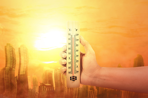 Hand holding thermometer with high temperature on the city with glowing sun background. Heatwave concept