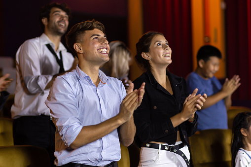 Amazed cheerful man and woman smiling and applauding while standing at seats in cinema hall after movie looking on stage