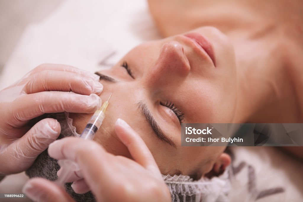 Beautiful young woman at cosmetology clinic Top view close up of a woman receiving filler injections in forehead. Professional cosmetologist injecting hyaluronic acid into skin of female client Botulinum Toxin Injection Stock Photo