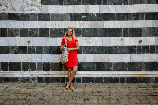 A front-view shot of a beautiful mature caucasian woman standing next to a patterned wall in Volterra, Italy, she is wearing a Red outfit.