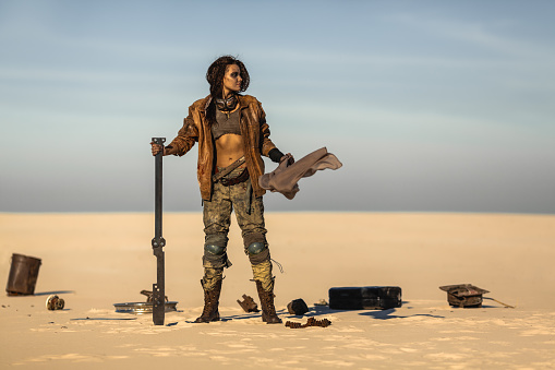 Post-apocalyptic woman with weapon outdoors. Young slim girl warrior in shabby clothes holding sword standing in a confident pose looking away. Nuclear post-apocalypse time. Life after doomsday concept. Desert and dead wasteland on the background.