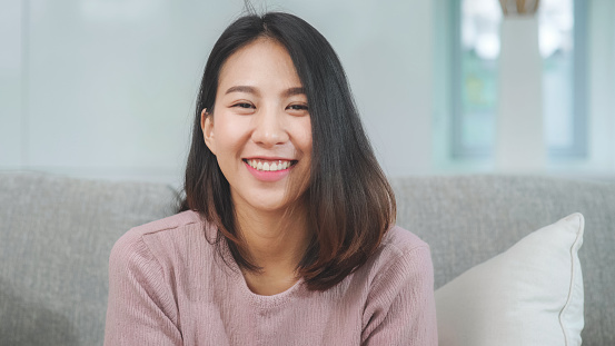 Teenager Asian woman feeling happy smiling and looking to camera while relax in living room at home. Lifestyle beautiful Asian young female using relax time at home concept.