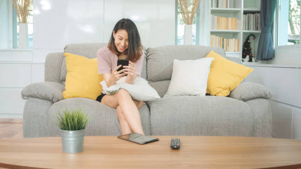 istock Young Asian woman using smartphone checking social media feeling happy smiling while lying on the sofa when relax in living room at home. Lifestyle latin and hispanic ethnicity women at house concept. 1188603109
