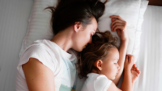 Family portrait of young mother is sleeping with her cute little daughter together on white linen in bed.