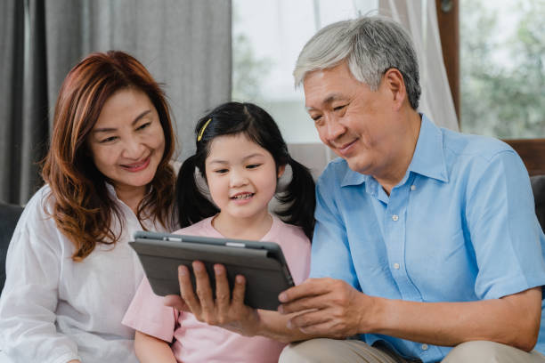 videochiamata per nonni asiatici e nipoti a casa. cinese anziano, nonno e nonna felici con la ragazza che usa la videochiamata del telefono cellulare che parla con papà e mamma sdraiati in salotto a casa. - ipad senior adult facebook sofa foto e immagini stock