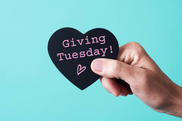 text giving tuesday in a heart-shaped sign the hand of a young caucasian man holding a black heart-shaped sign, with the text giving tuesday written in it, on a pink background giving tuesday stock pictures, royalty-free photos & images