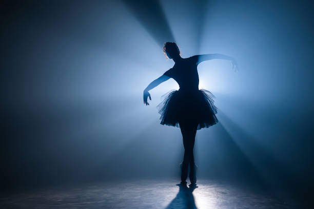 검은 투투 드레스를 입은 발레리나는 마술 같은 푸른 빛과 연기로 무대에서 춤을 추고 있습니다. 발레 슈즈 포인테로 젊은 매력적인 댄서의 실루엣이 어둡게 공연. 공간을 복사합니다. - 여성댄서 뉴스 사진 이미지