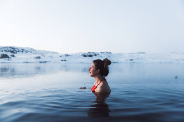 アイスランドの美しい雪に覆われた山々の景色を眺めた温泉プールで泳ぐ女性 - 凍っている水 ストックフォトと画像