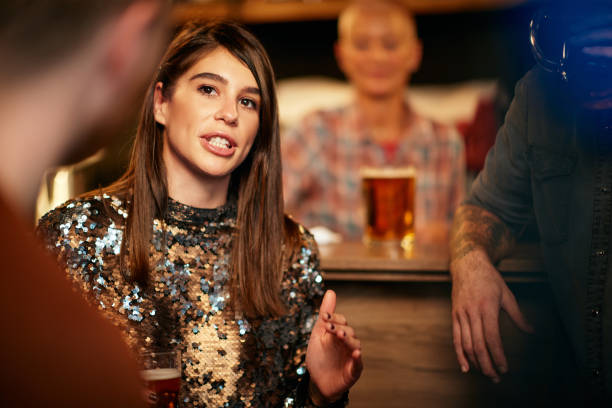 Beautiful caucasian smiling stylish woman  holding beer and chatting with friends. In background is bar counter. Pub interior.