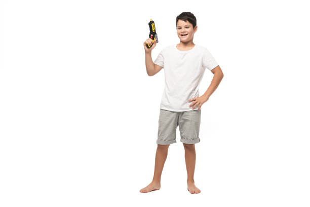 腰に手を置き、白い背景にカメラで微笑みながらおもちゃの銃を持つ陽気な少年 - toy gun ストックフォトと画像