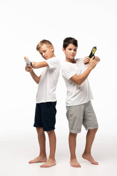 due fratelli in piedi back to back, tenendo le pistole giocattolo e guardando la fotocamera su sfondo bianco - toy gun foto e immagini stock