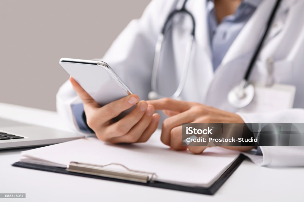 Male doctor using smartphone at workplace, making online consultation Consultation Online. Unrecognizable male doctor using smartphone at workplace, messaging with his patient, prescribing treatment Doctor Stock Photo