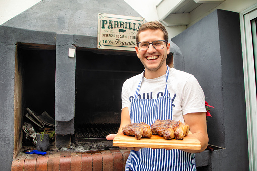 Millennial Argentinian cook presenting roasted rib eye and strip steaks fresh from his parilla BBQ seen in the background. These are ojo de bife and bife de chorizo cuts. He smiles warmly at the camera.