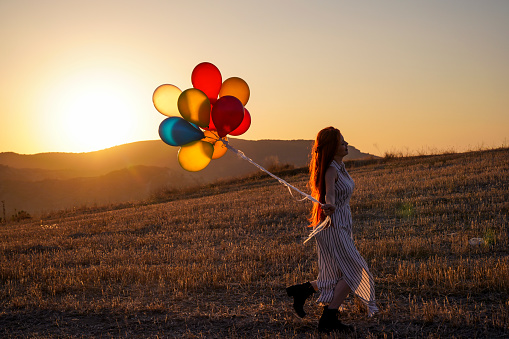 Happy cheerful girl with balloons running across meadow at sunset on nature in summer