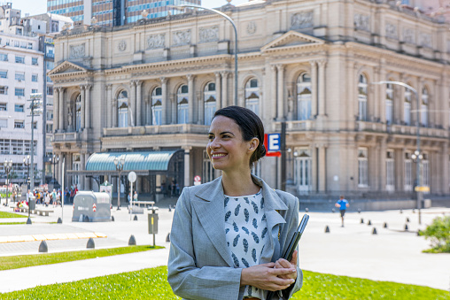 Portrait of a beautiful young businesswoman smiling holding note pads in a public park at Buenos Aires, Argentina. At the background the facade of the Colon Theater building.