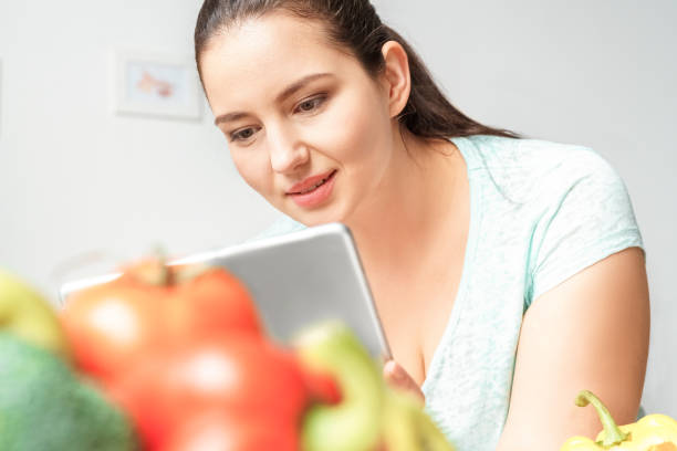istock Body Care. Chubby girl leaning on table with vegetables in kitchen browsing internet on digital tablet concentrated close-up 1188759482