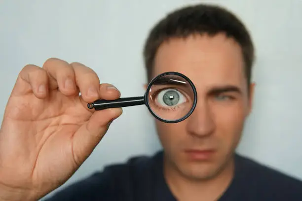 Potrait of a man holding magnifying glass and looking through it srewing up one eye. Funny view to magnified man's glass through magnifier. Man uses loupe to look intently to the some object. Macro