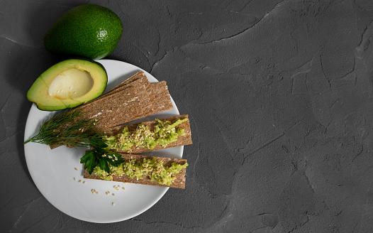 Top view crispbreads with mashed avocado sprinkled sesame seed and slice fresh avocado on plate with empty space for text with black concrete background
