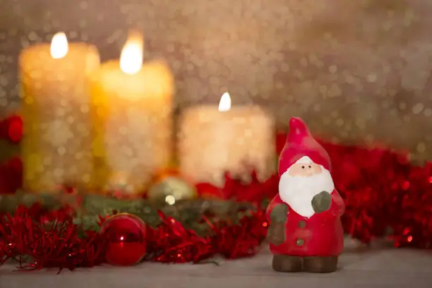 Photo of The warmth of the Christmas concept: a Santa Claus statuette in foreground in a rustic setting with with three candles lit in bokeh effect, gold and red bright baubles, snow effect