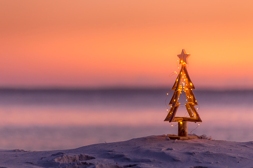 A coastal summer Christmas in Australia.  A driftwood Christmas tree decorated with fairy lights on the beach in summer sunrise or sunset