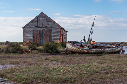 Thornham, Norfolk, England, UK - April 24, 2019: An old stone barn in Thornham Old Harbour with a wooden sailing boat next to it