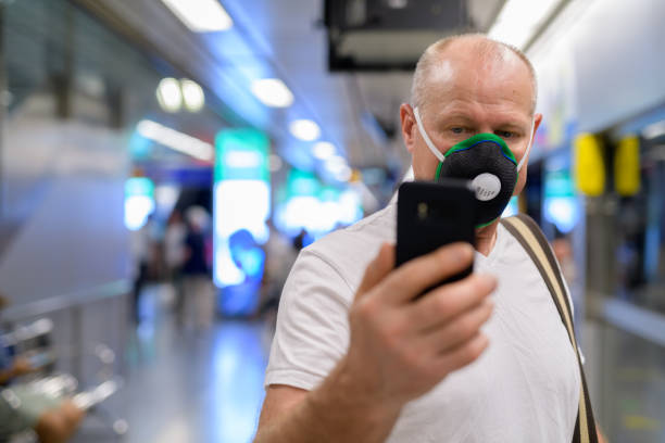 Senior man using face mask to protect from pollution smog in city while holding mobile phone at train station