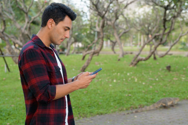 istock Profile view of young handsome Hispanic hipster man using phone at the park 1188845416