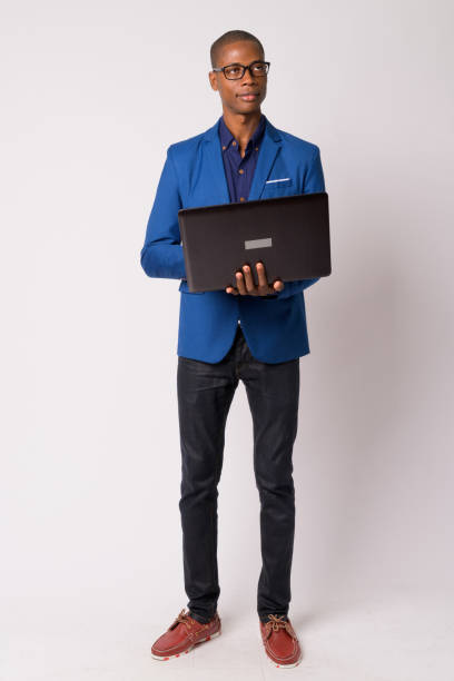 Full body shot of young bald African businessman thinking while using laptop Studio shot of young handsome bald African businessman in suit with eyeglasses against white background black nerd stock pictures, royalty-free photos & images