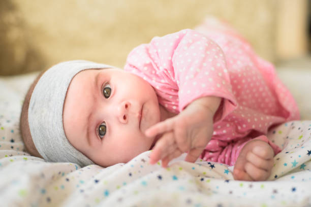 carina bambina di 6 mesi su un letto sulla pancia con la testa alta che guarda in macchina fotografica con i suoi grandi occhi. luce naturale. - sdraiato su un fianco foto e immagini stock