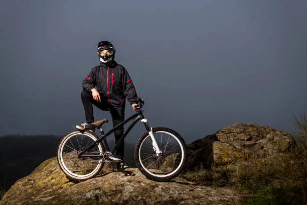 The rider in the full-face helmet and full protective equipment on the mtb bike stands on a rock. Downhill, MTB.