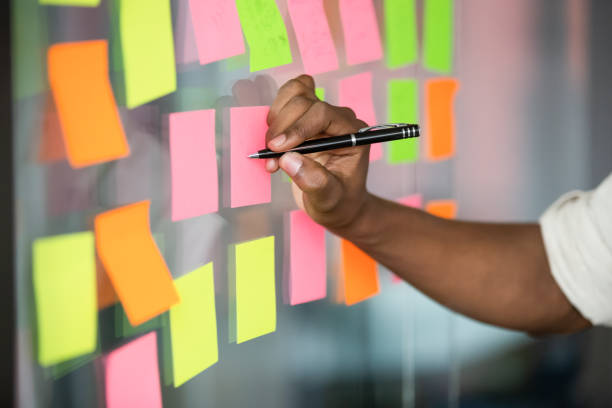 Close up african american manager writing notes on kanban board. Close up african american manager writing notes on colorful sticky notes on kanban board. Mixed race employee managing project workflow, scheduling tasks or planning strategy on glass wall at office. agility stock pictures, royalty-free photos & images