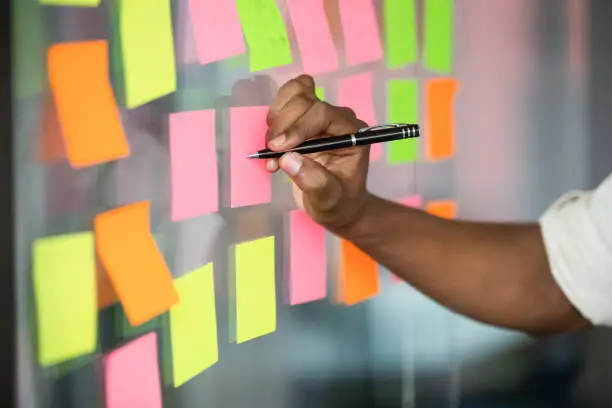Close up african american manager writing notes on colorful sticky notes on kanban board. Mixed race employee managing project workflow, scheduling tasks or planning strategy on glass wall at office.