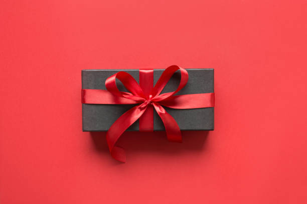 Black gift box wrapped with red ribbon, bow on red surface. Top view. Space for text.