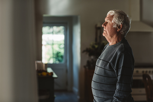 Senior man looking sad and lost in thought while standing alone in his home looking out through a window