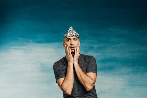 A man in a foil hat scaredly grabs his hands with his hands wide open his eyes and mouth on a blue background. A man in a foil hat scaredly grabs his hands with his hands wide open his eyes and mouth on a blue background. tin foil hat stock pictures, royalty-free photos & images