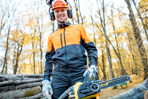 Portrait of a handsome professional lumberjack in protective workwear with a chainsaw near the wooden logs in the forest