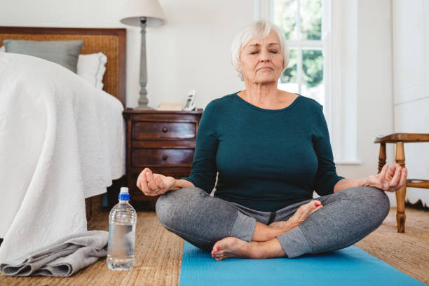 Active senior woman in the lotus pose while practicing yoga Active senior woman sitting on a mat on her bedroom floor meditating in the lotus pose while practicing yoga in the morning grey hair on floor stock pictures, royalty-free photos & images